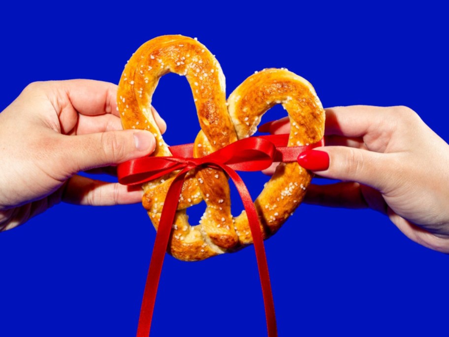 two hands holding a heart shaped pretzel in front of a blue background