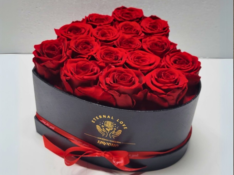 a black heart shaped box with gold lettering and a red ribbon around it filled with red roses 