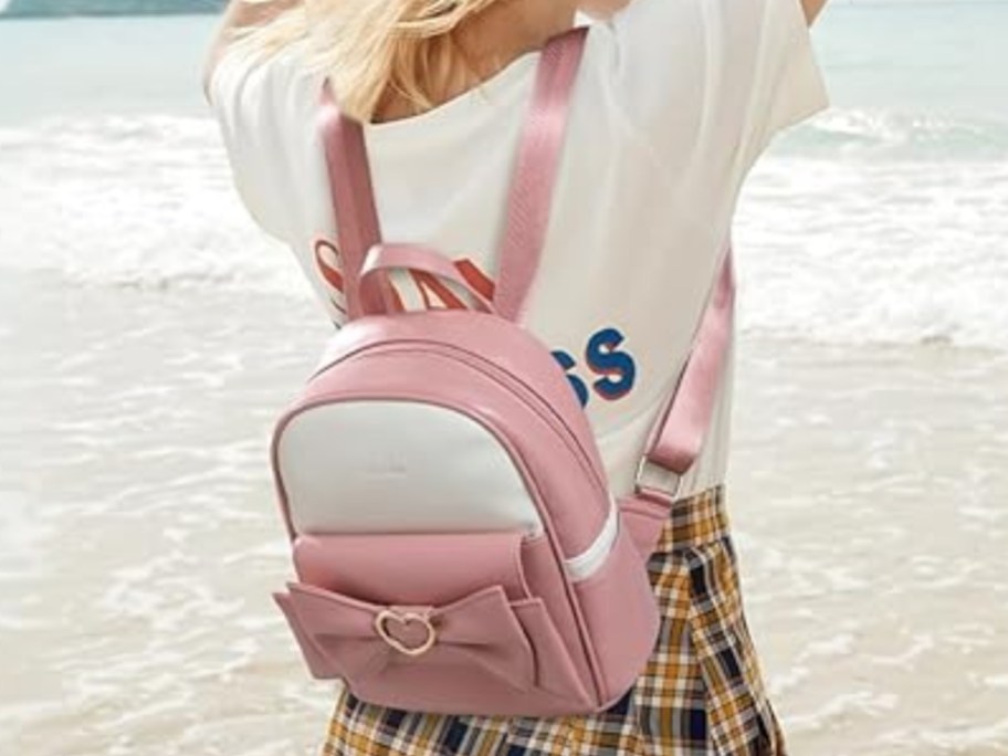 woman standing on the beach, she's wearing a tshirt and plaid skirt and has a pink and white mini backpack purse on her back that has a bow on it