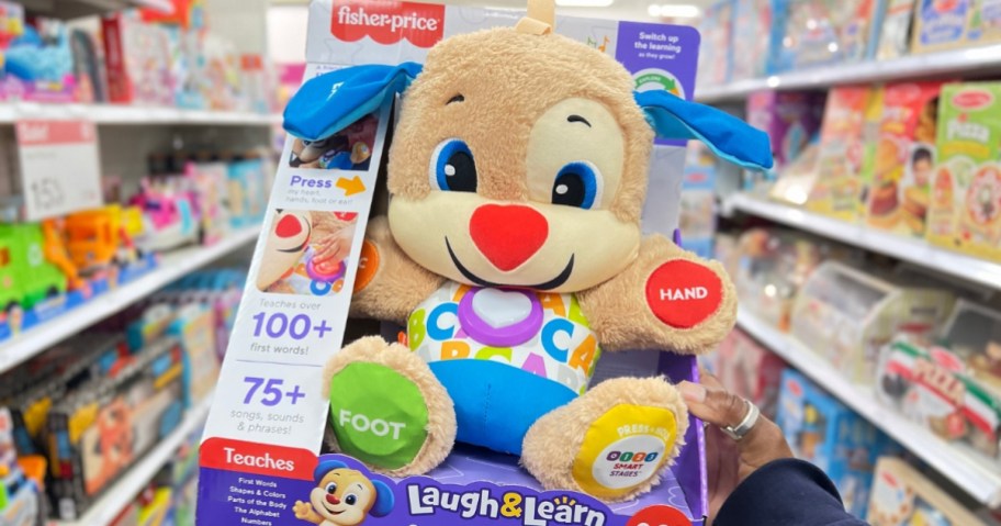 hand holding a Fisher-Price Laugh & Learn Puppy toy that's in the packaging box it comes in, a store aisle is behind it