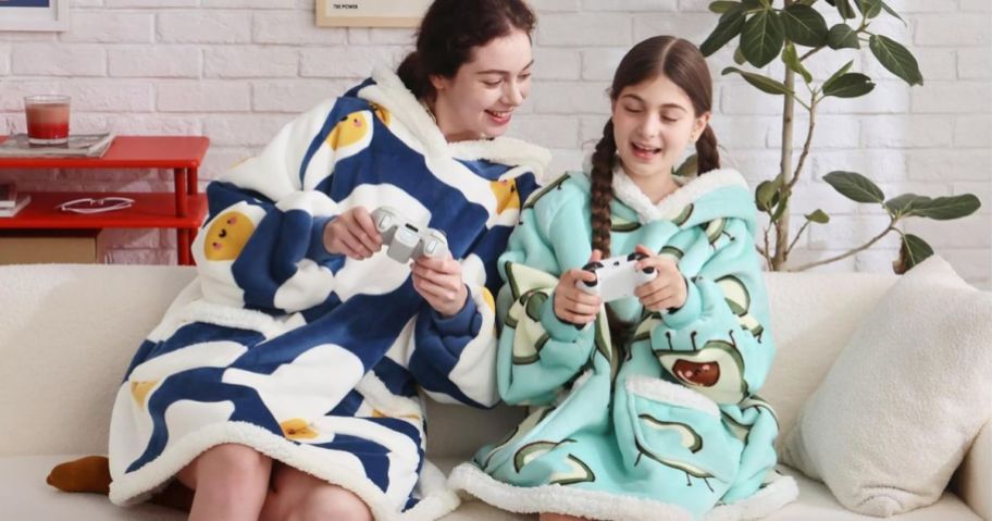 A woman and a gil sitting on a couch playing video games wearing Bedsure wearable blankey hoodies