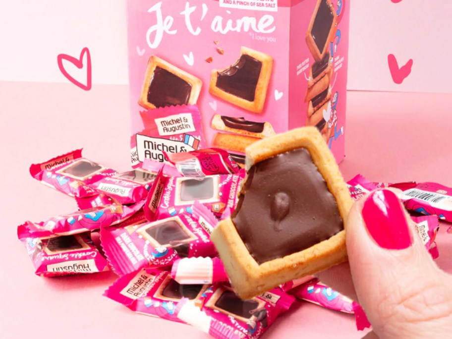 hand holding up a square Michel et Augustin Cookie in front of a pile of pink individually wrapped cookies