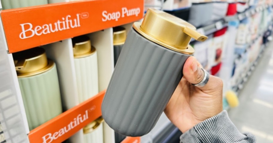 hand holding a soap pump that is a grey ribbed design with a gold top and pump, you can see a display with more colors of the soap pump in a store aisle behind it