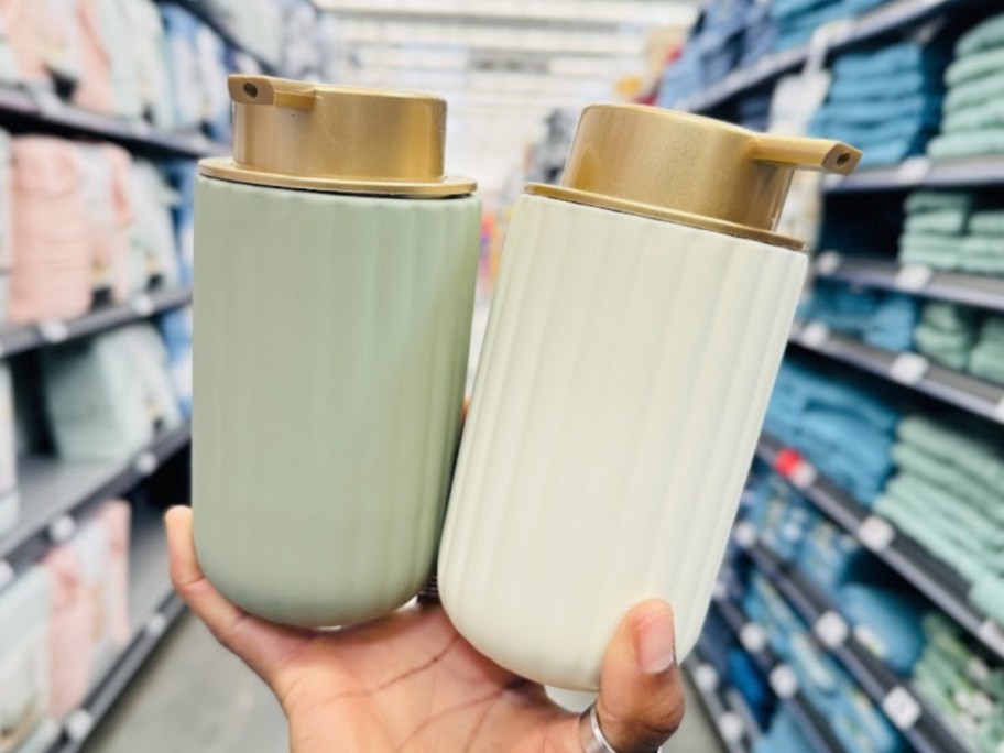 hand holding 2 soap pumps, one is light green ribbed and one is off white ribbed, both have a flat gold color soap pump top