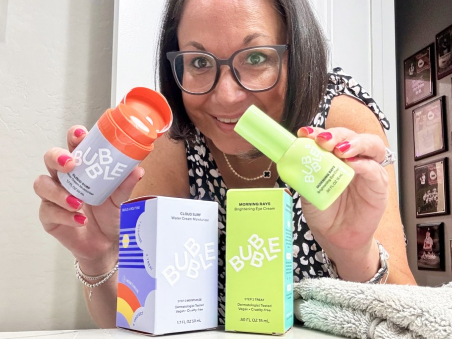 woman holding up two Bubble Skincare <a href='https://hadesproducts.com/products' target='_blank' rel='follow'>products</a> in bathroom” width=”912″ height=”684″></p>
<div class=