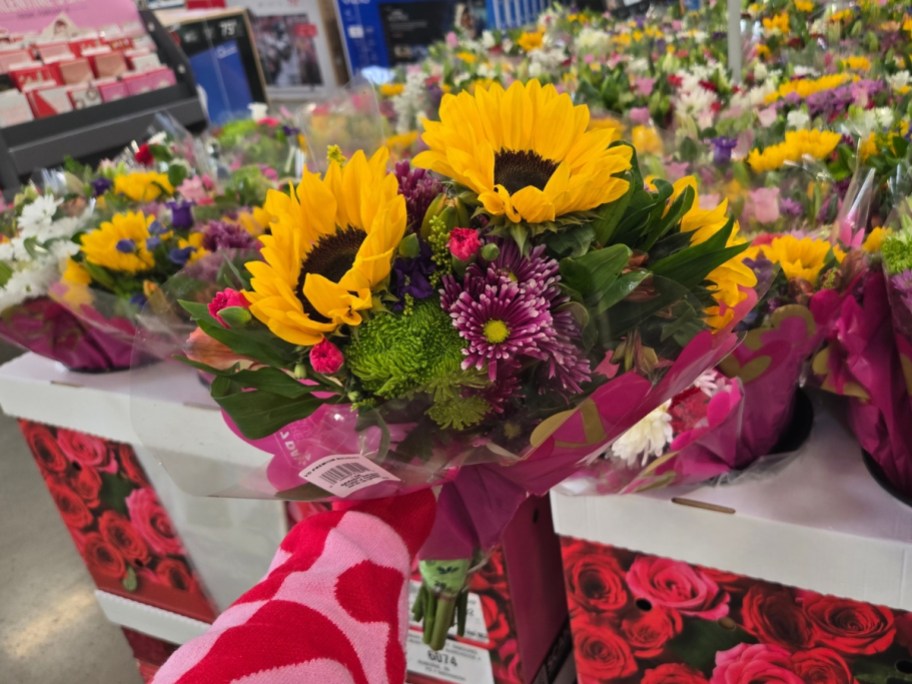 hand holding a bouquet of flowers with sunflowers, and pink and purple flowers wrapped in plastic - a display with more flower bouquets is behind it