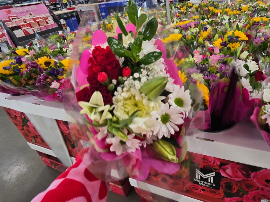 hand holding a bouquet of flowers with white lilies, white daisies, and red roses wrapped in plastic - a display with more flower bouquets is behind it