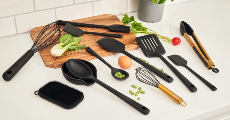 a set of black and gold kitchen utensils on a kitchen counter, wood cutting board and foods around them