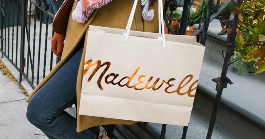 woman standing outside leaning against a wrought iron fence wearing jeans and a tan coat and plaid scarf holding a cream and gold shopping bag that says "Madewell"