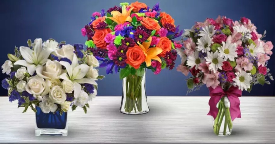 3 vases of flowers,one has white lilies, white roses, and purple flowers, one has orange roses and pin, blue, peach, and purple flowers, the other has white, pink, and purple daisies and other flowers