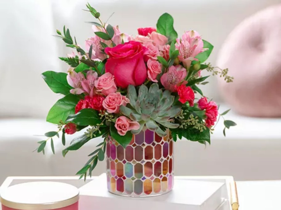 a mosaic glass vase with dark pink roses and pink and purple flowers and green succulents sitting on a coffee table