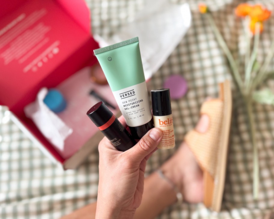 hand holding face cream, blush stick, and serum above red box