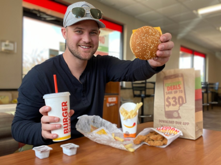 man holding up hamburger with soda and other fast food in restaurant