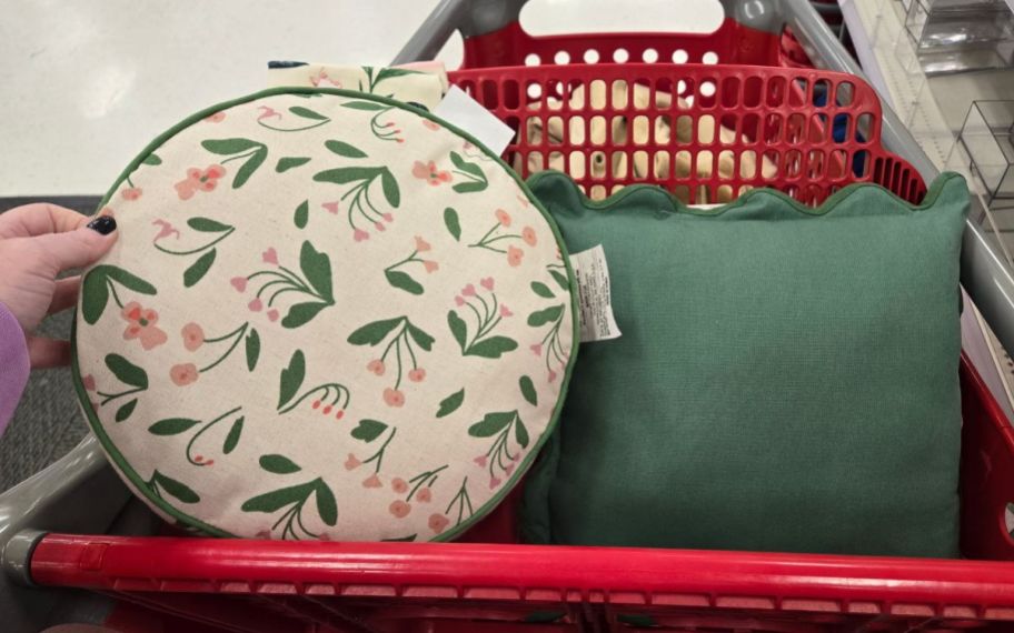 a womans hand holding a round floral print throw pillow next to a green one in a shopping cart.