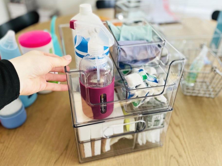 hand touching stack of storage bins filled with cleaning supplies