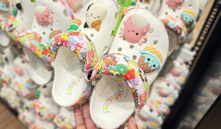 hand holding a pair of Squishamllows Crocs clog style shoes, with a display of more shoes behind them