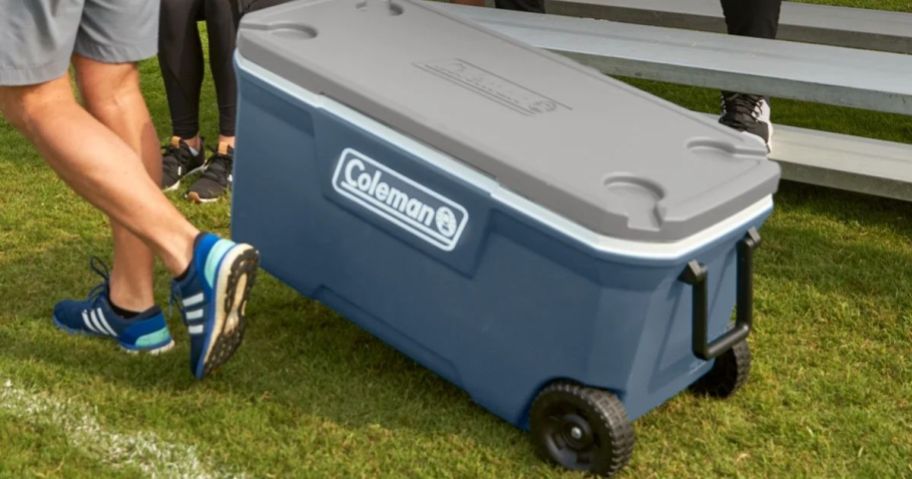 man pulling a large blue Coleman wheeled cooler by the handle across the grass