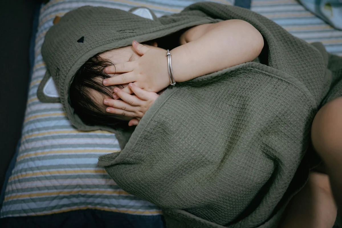 Child hides face under hooded towel on striped bed - infant child care - Baby gear and nursery furniture discount offers