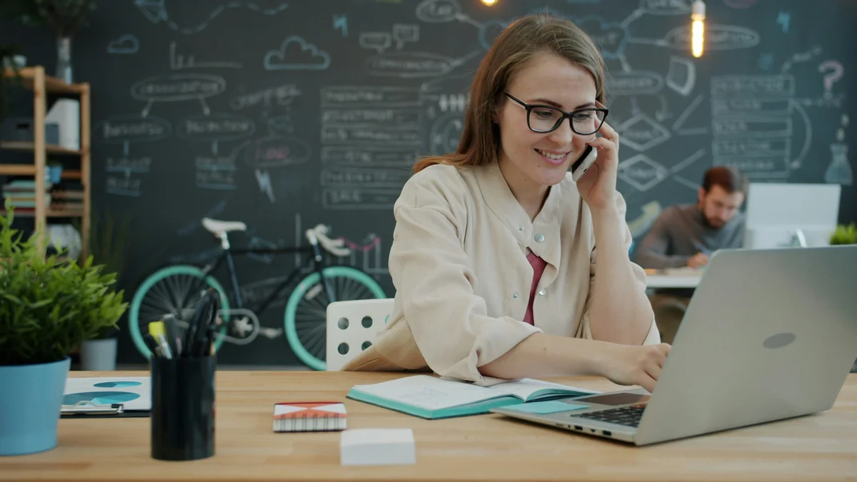 Businesswoman is busy with phone call talking and using laptop working with laptop computer in open space office, colleagues are in background. - exclusive software license discounts for small businesses