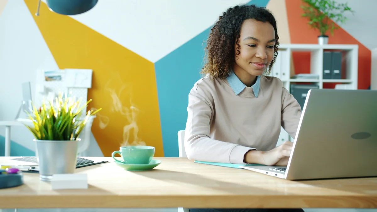Cinemagraph loop of beautiful Afro-American girl using laptop in office with hot drink on table, steam is going up from cup. People and workplace concept. - exclusive software license discounts for small businesses