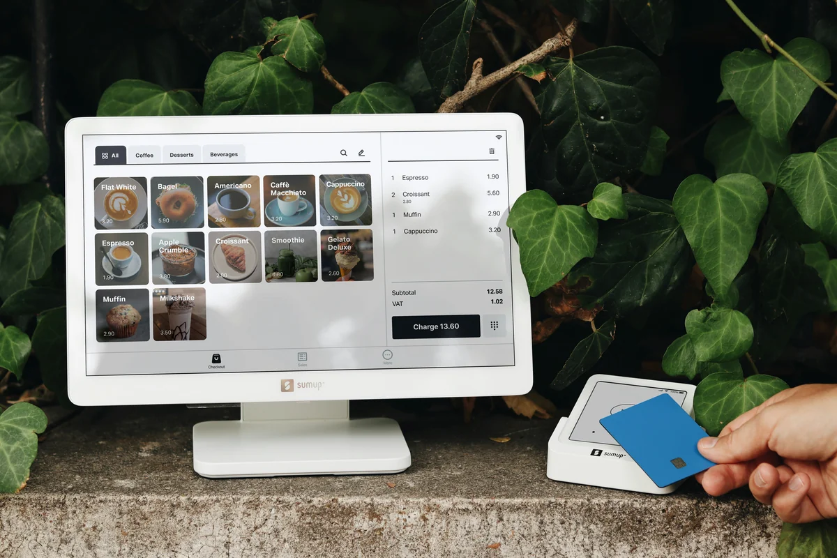 A SumUp POS system is set up in an outdoor, nature-inspired setting, with a user making a contactless payment using a blue card. The screen displays a cafe menu with items like coffee, croissants, and desserts, creating a vibrant and inviting retail experience. The clean design of the SumUp system, combined with the lush greenery surrounding it, enhances the natural, eco-friendly aesthetic of this modern payment solution, ideal for cafes, coffee shops, and small businesses. - exclusive software license discounts for small businesses