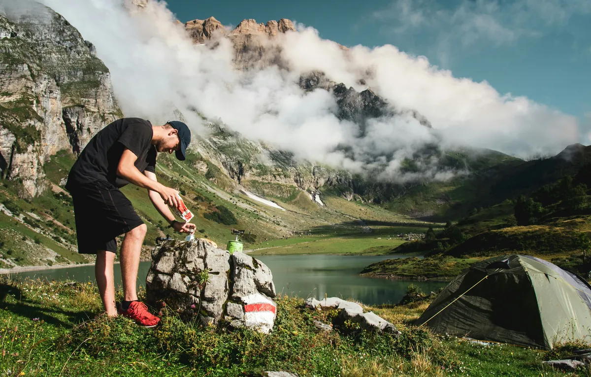 man in black t-shirt and gray shorts standing on rock near lake during daytime - outdoor camping gear deals and hiking equipment sales