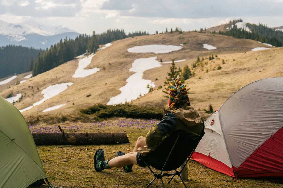 a person sitting on a chair outside by a tent and a lake - outdoor camping gear deals and hiking equipment sales