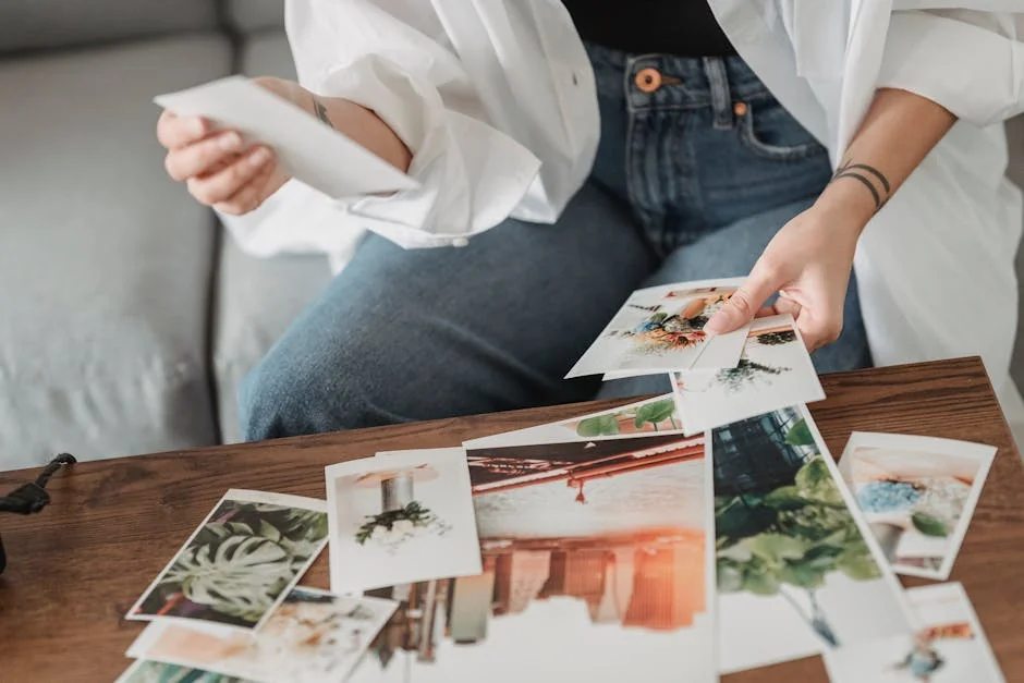 Crop unrecognizable tattooed female sitting on sofa at desk with assorted photos of flowers and town in house room - photo printing coupons and personalized gift deals