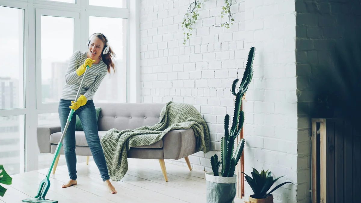 Pretty blond girl is washing floor with flat plastic mop and listening to music in headphones, dancing and singing during routine housework in beautiful apartment. - spring cleaning supply coupons and household deals